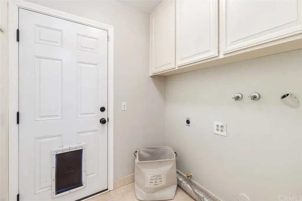 Laundry room with white door, beige walls, cabinets, water connections, trash bin, and pet door.