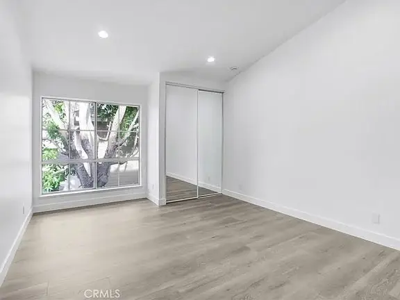 Empty room with white walls, large window with greenery, mirrored closet door, and light wood floor