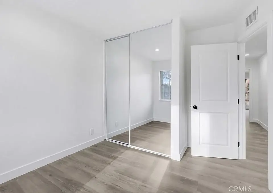 Empty room with light wood floor, white walls, mirrored closet door, and open door to hallway