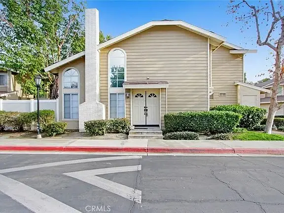 Two-story house with light brown exterior, white trim, chimney, arched windows, doors, bushes, and trees