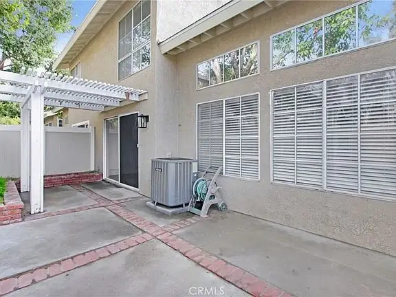 Patio with concrete and brick paving, beige house with large windows, white structure, air conditioning unit, and hose reel