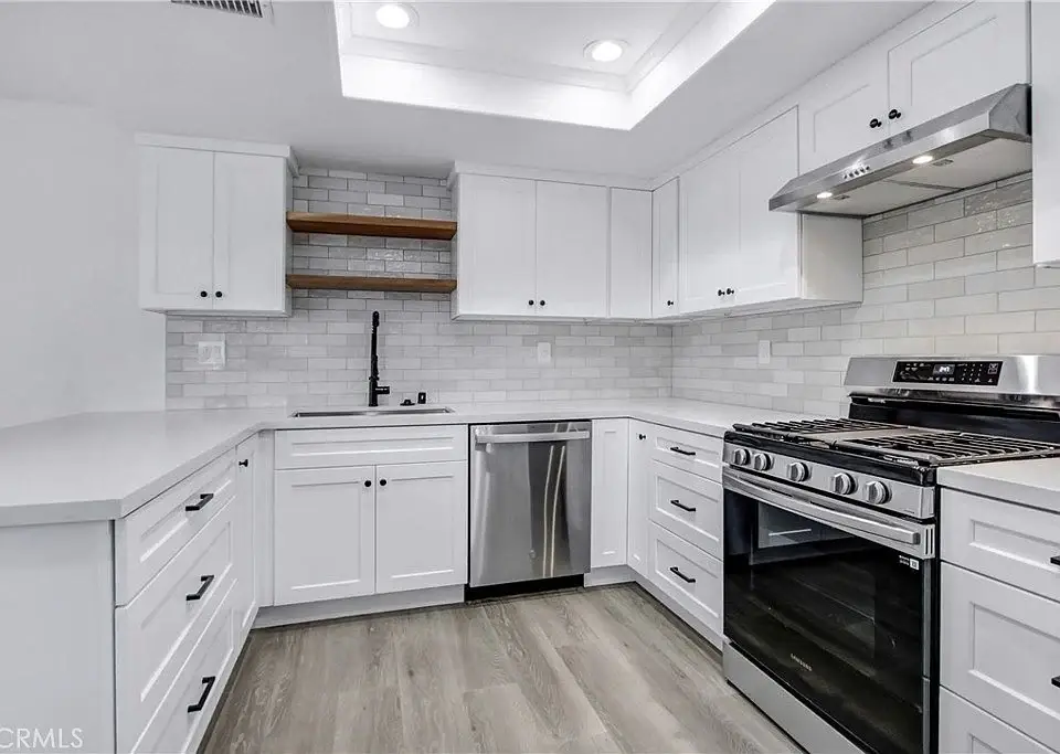 Kitchen with white cabinets, gas stove, dishwasher, light countertops, grey backsplash, and wooden shelves