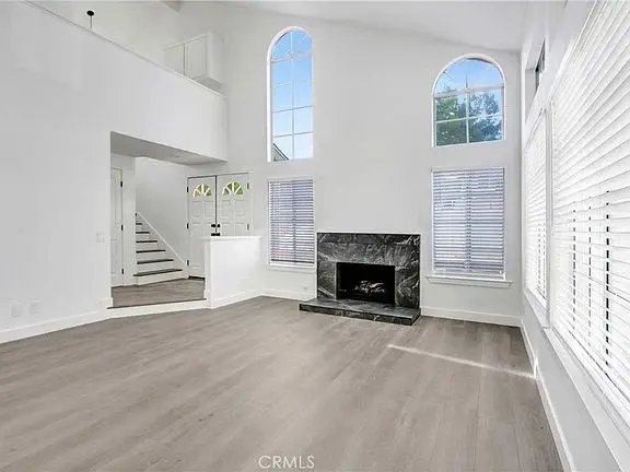 Living room with large windows, white walls, light wood floor, black stone fireplace, and staircase visible