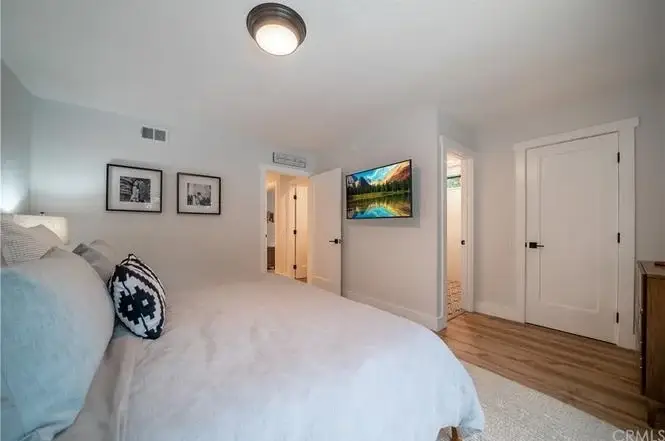 Bedroom with bed, white linens, decorative pillows, wall-mounted TV, framed photos, light walls and floor, and doorway