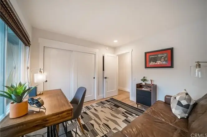 Small room with desk, chair, brown leather sofa, area rug, window with blinds, framed artwork, and plant