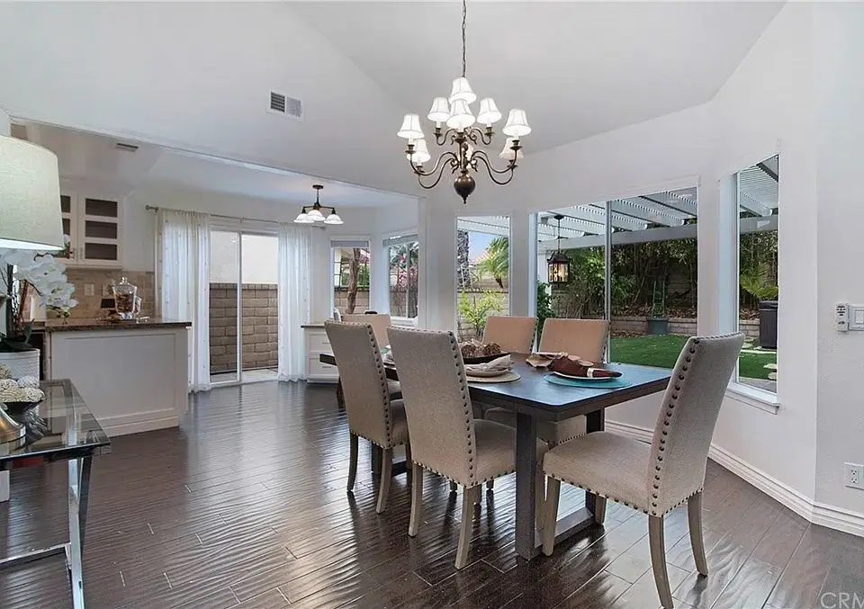 Dining area with table, high back chairs, chandelier, big windows, wood floor, and view of greenery.