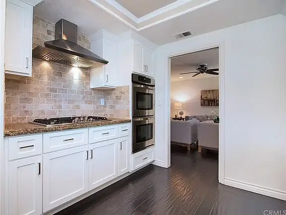 Kitchen with white cabinets, granite counter, stainless double oven, range hood, tile backsplash, and open doorway.