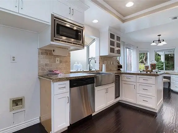 Kitchen with white cabinets, dishwasher, microwave above stove, sink window, stone counter, and wood floor.