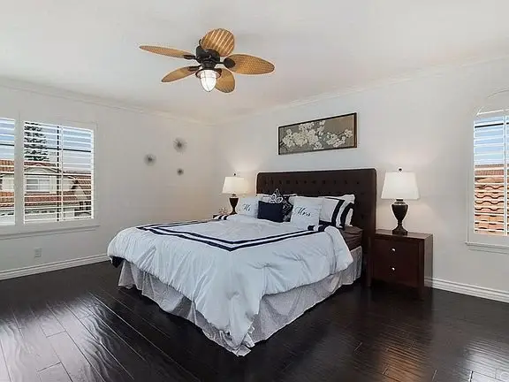 Bedroom with large bed, navy and white bedding, wood headboard, lamps, ceiling fan, shutters, and wood floor.