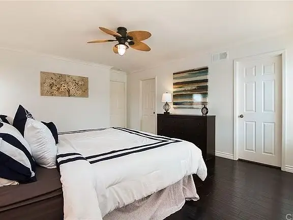 Bedroom with large bed, white and navy bedding, dresser, wall art, ceiling fan, and door.