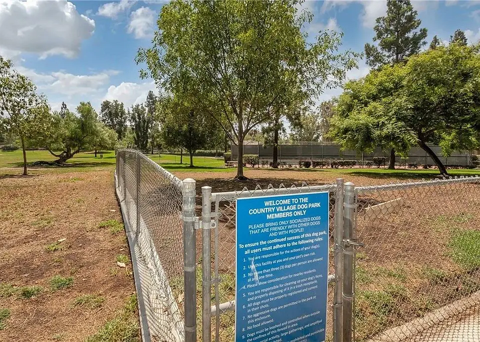 Dog park with sign for Country Village Dog Park, fenced area, grass, and trees.
