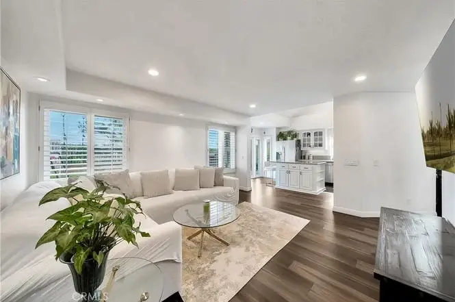 Living room with light sofa, round glass coffee table, rug, big windows, and kitchen in back.