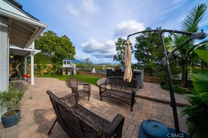Backyard patio of house California real estate with wicker chairs, umbrella, grass, trees, and play ground