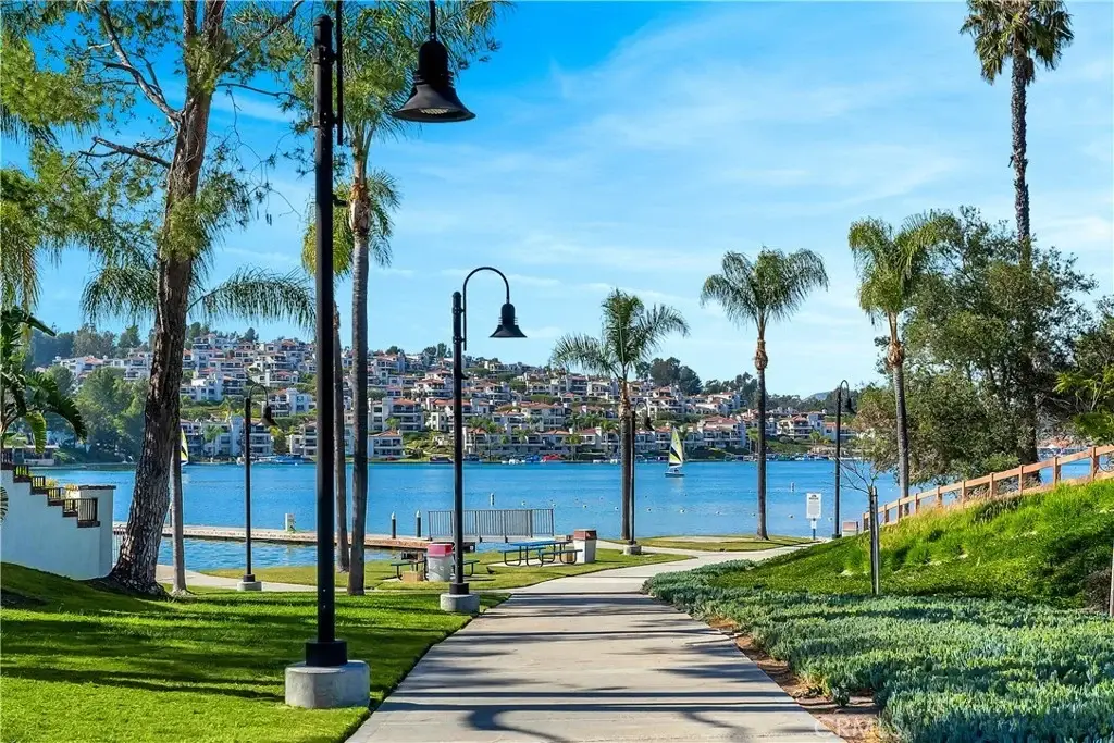 Pathway with black lampposts and palm trees leading to lake with sailboats and houses on hill.