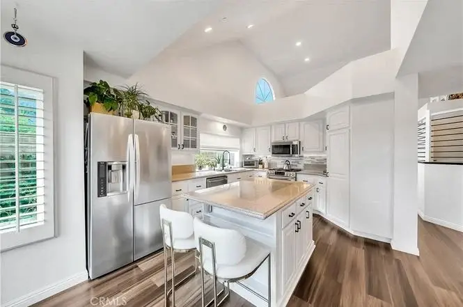 Kitchen with white cabinets, stainless appliances, large island with seating, windows, and high ceiling.