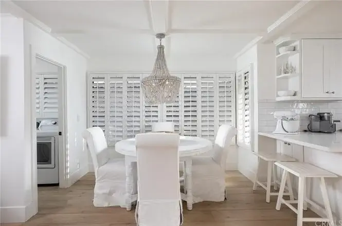 Dining room with round white table, white chairs, chandelier, wood floor, window shutters, and bar stools.