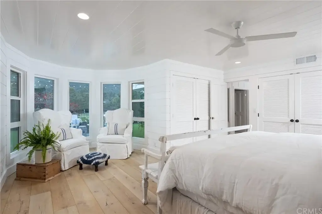 Bedroom with bed, two white chairs by bay window, ceiling fan, white walls, and wood floor.