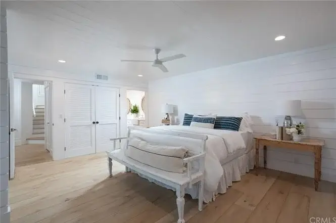Bedroom with king bed, white linens, blue striped pillows, wood bench, ceiling fan, wood floor, and doorway.