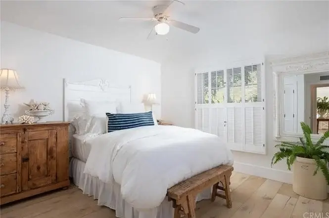 Bedroom with white bed, blue striped pillows, wood dresser, ceiling fan, plants, shutters, and wood floor.