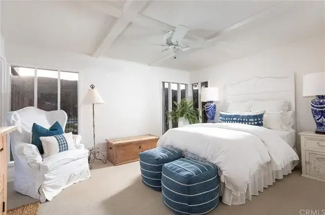 Bedroom with white bed, blue pillows, striped ottomans, wood chest, lamp, plants, and windows.