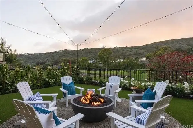 Backyard of California real estate house with fire pit, Adirondack chairs, blue pillows, string lights, and hillside.