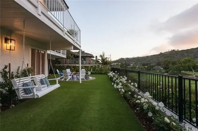 Backyard of house California real estate with artificial grass, swing set, benches, hedges, and flowers.