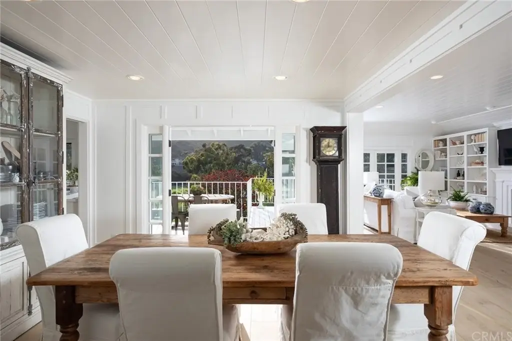 Dining area with wood table, white chairs, cabinet, clock, and large windows.