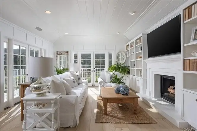 Living room with white sofa, wood coffee table, fireplace with wall TV, shelves, plants, and large windows.