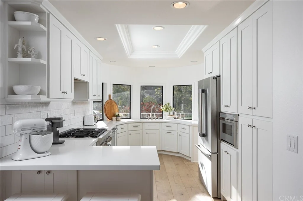 Kitchen with white cabinets, island seating, stainless steel appliances, wood floor, and large windows.