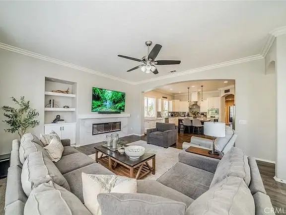 Living room with gray sectional sofa, coffee table, flat-screen TV, ceiling fan, and open kitchen in background