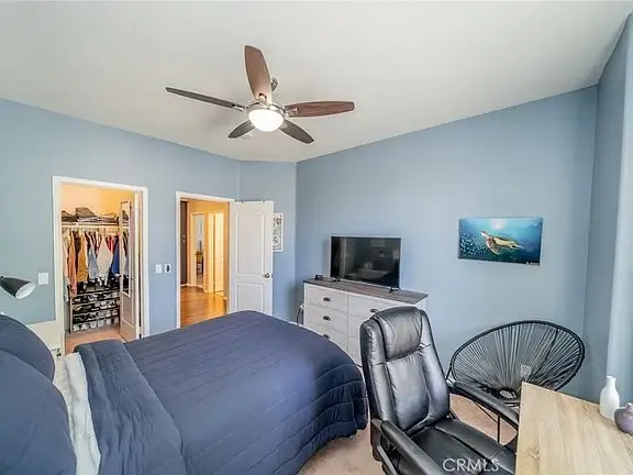 Bedroom with blue wall, bed with navy comforter, office chair, dresser with TV, ceiling fan, and open closet