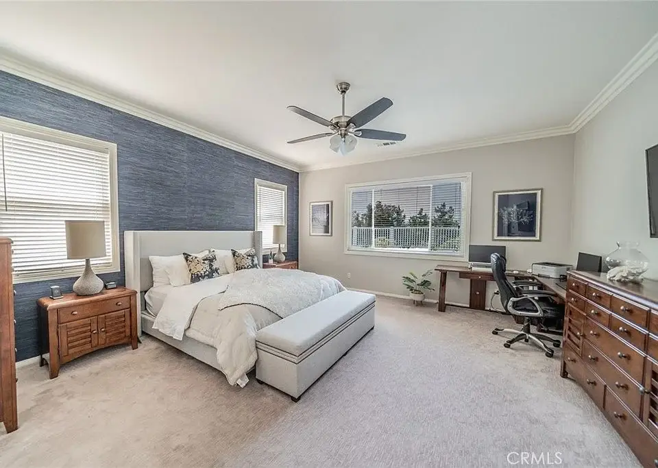 Bedroom with bed, decorative pillows, ceiling fan, desk, blue walls, windows, and carpet