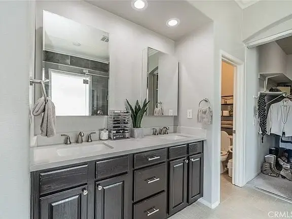 Bathroom with dual sinks, gray cabinets, large mirrors, decor, and door to closet