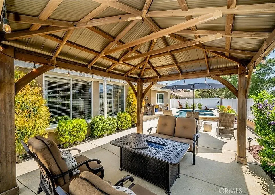 Covered patio with table, chairs, greenery, and swimming pool in background