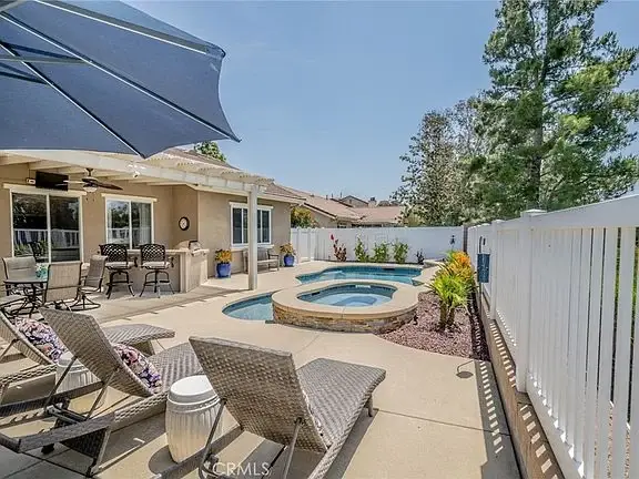 Backyard patio with lounge chairs, umbrella, hot tub, and colorful plants along white fence