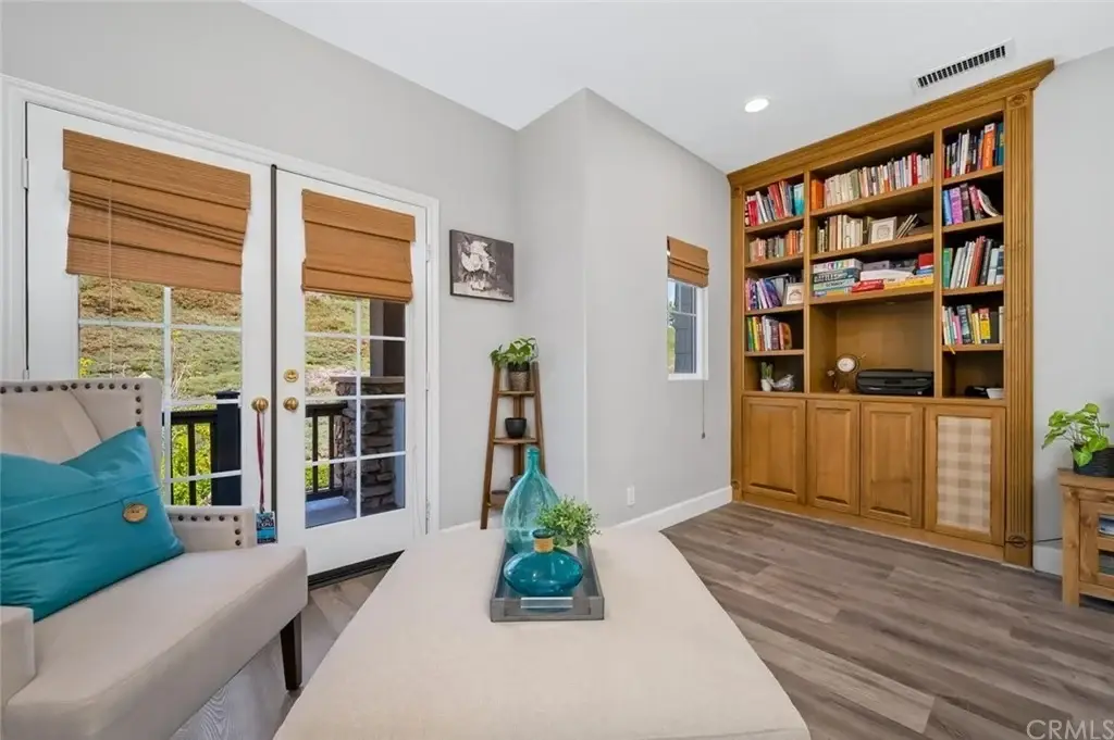 Living room with beige chair, light coffee table, wood shelves with books, window doors, and plants.
