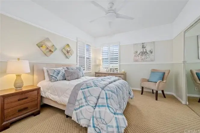 Bedroom with queen bed, blue and white quilt, wall art, desk, beige chair, and window shutters.