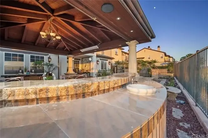Patio of house California real estate with stone counter, outdoor seating, lights, plants, and trees.