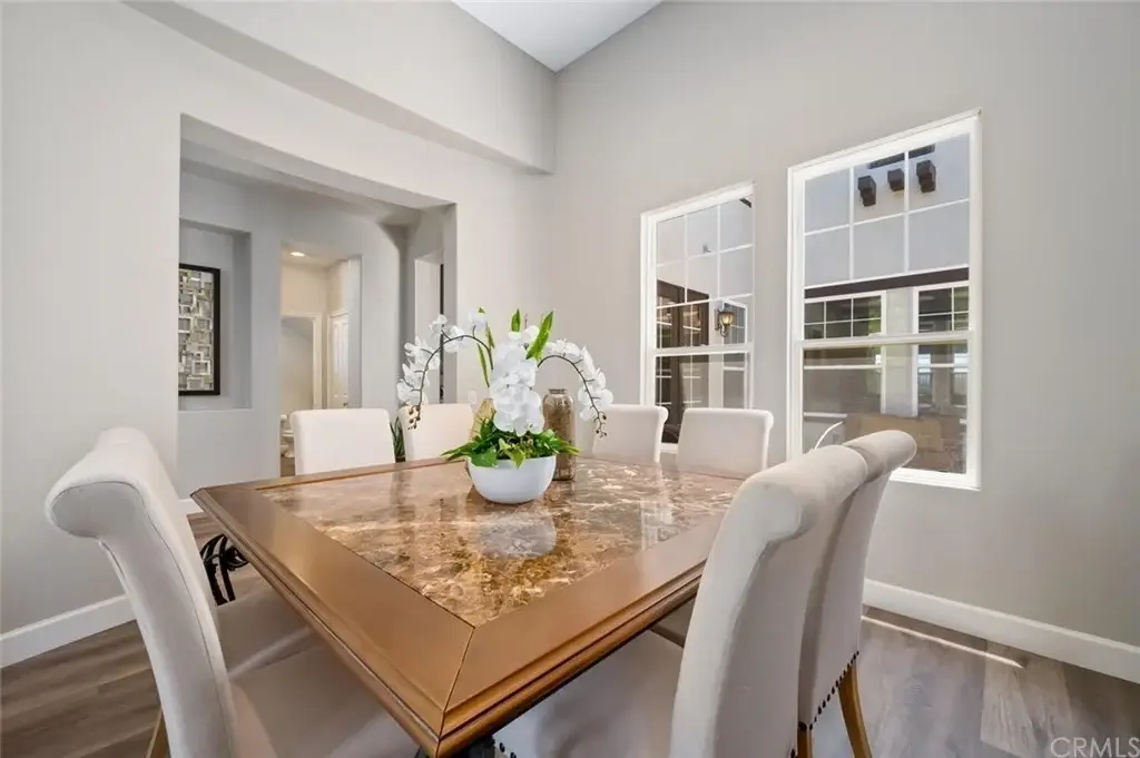 Dining room with wooden table, chairs, white orchids centerpiece, light walls, and windows.
