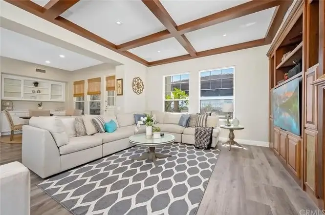 Living room with light couch, coffee table, geometric rug, coffered ceiling, wall TV cabinet, plants, and large windows.