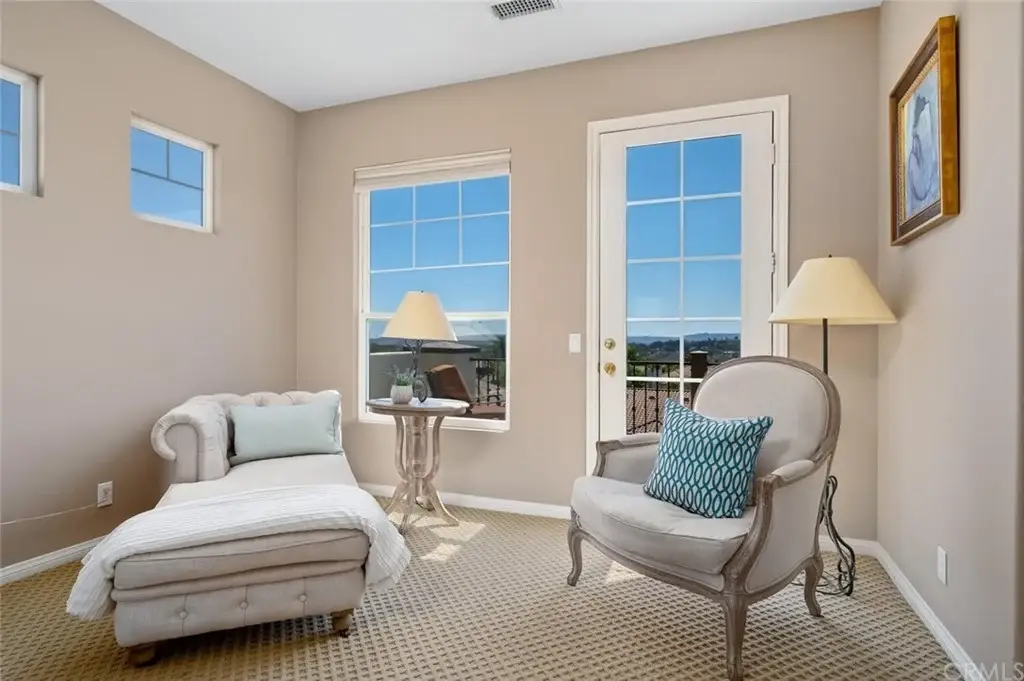 Room with chaise lounge, side table with lamp, chair with blue pillow, and large windows.