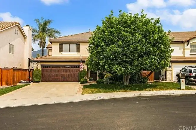 Two-story California real estate house with brown garage door, greenery, large front yard tree, on a corner lot.