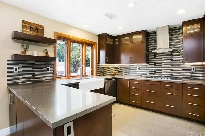 Kitchen with dark wood cabinets, large sink under window, striped tile backsplash, sleek countertop, and shelves with decorative items.