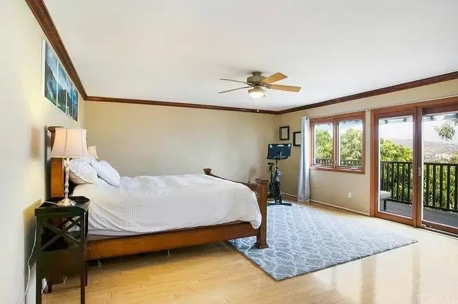 bedroom with wooden bed, white bedding, wooden floor, ceiling fan, large windows, and decorative area rug.