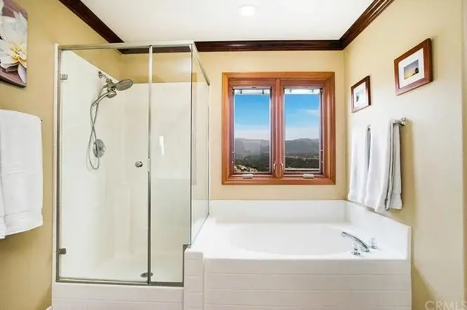 Bathroom with glass shower, white tub, windows with scenic view, framed pictures, and towels.