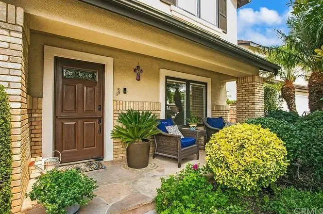 Porch with wooden front door, decorative brickwork, potted plants, and seating with blue cushions.