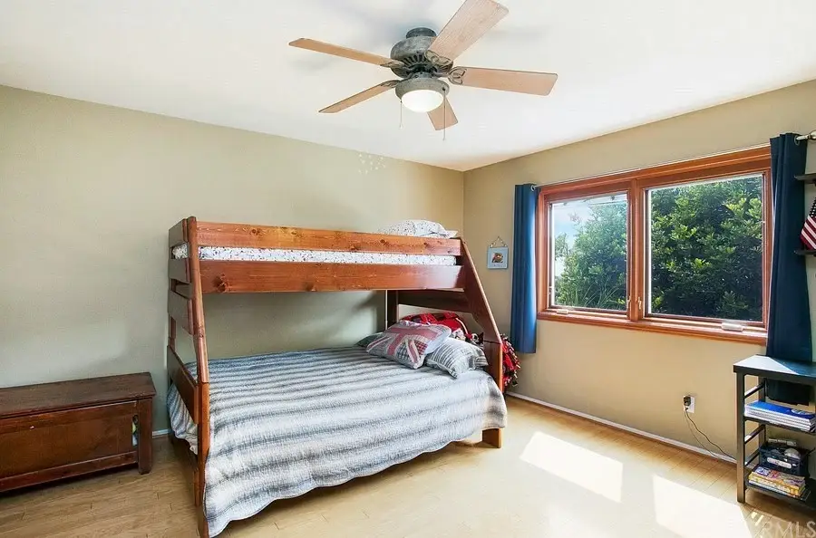 Bedroom with wooden bunk bed, lower bed with blankets, ceiling fan, large windows with blue curtains, wooden dresser, and shelf unit.