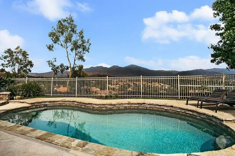 Swimming pool with stone border, lounge chairs, trees, mountains, and fenced area.