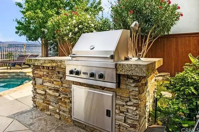 Outdoor stainless steel grill on stone countertop near pool and greenery.