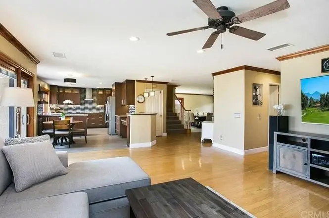 Living room with gray sectional sofa, wooden floor, ceiling fan, TV, and open kitchen with dark cabinets and dining area.